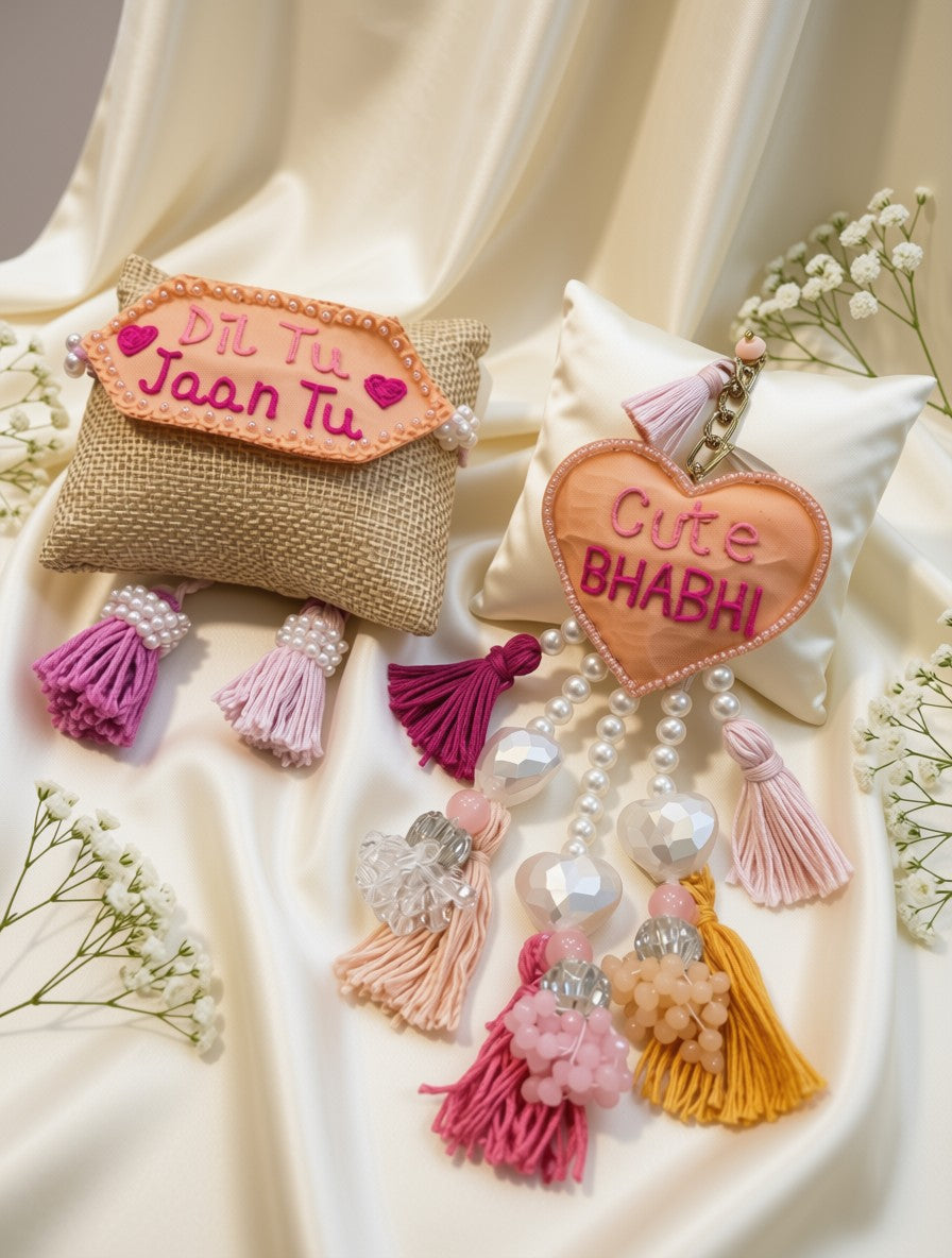 Two embroidered fabric keepsake pouches with tassels and pearl-like beads, styled on cream satin fabric with baby's breath flowers. One pouch is tan burlap with a peach embroidered label reading 'Dil Tu Jaan Tu' in pink, and the other is white with a peach embroidered label reading 'Cute BHABHI' in pink.