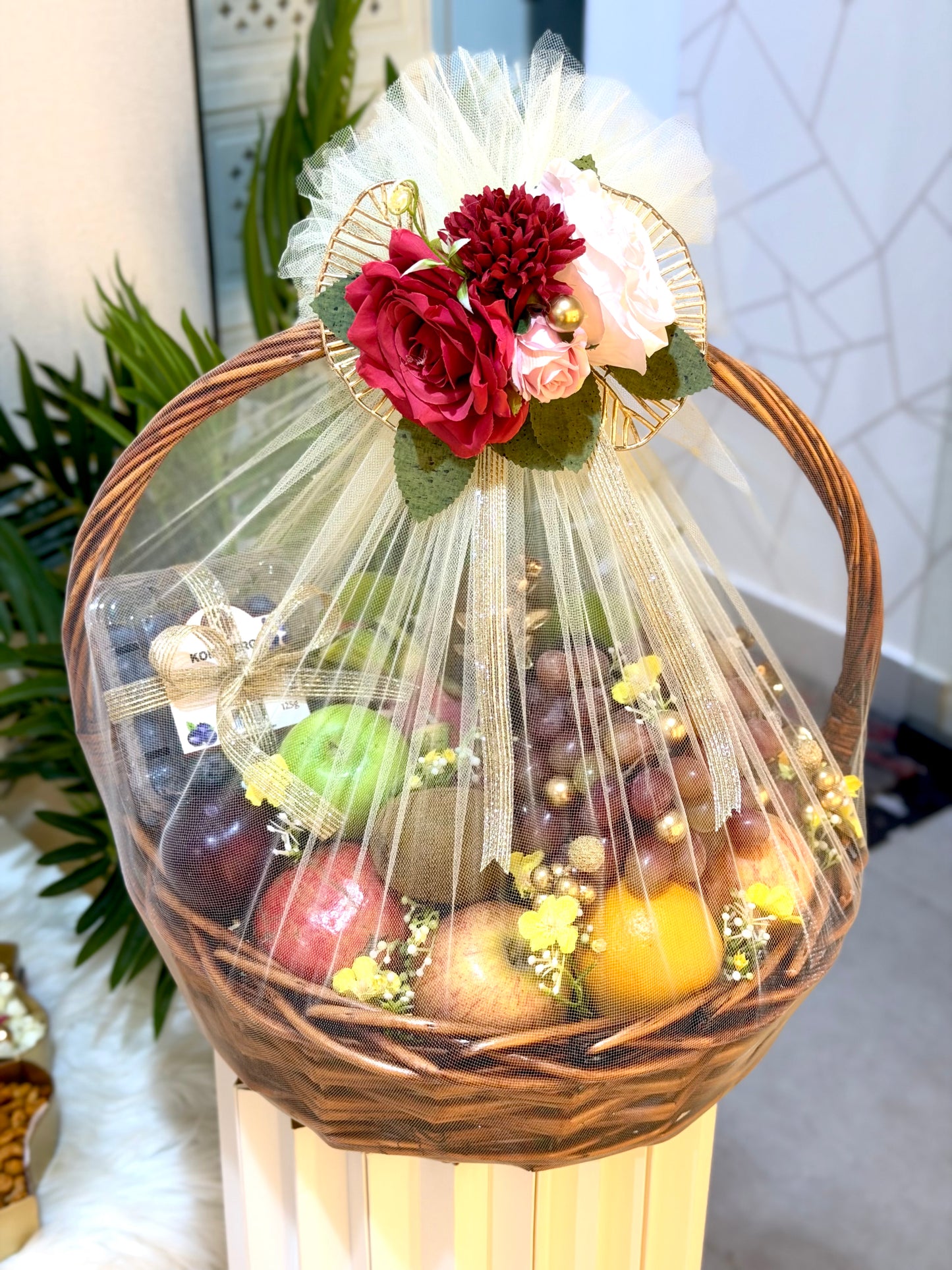 A wicker gift basket filled with assorted fruits, wrapped in clear cellophane and decorated with a white tulle bow, red and pink flowers, and green leaves, displayed on a pedestal in an indoor setting.