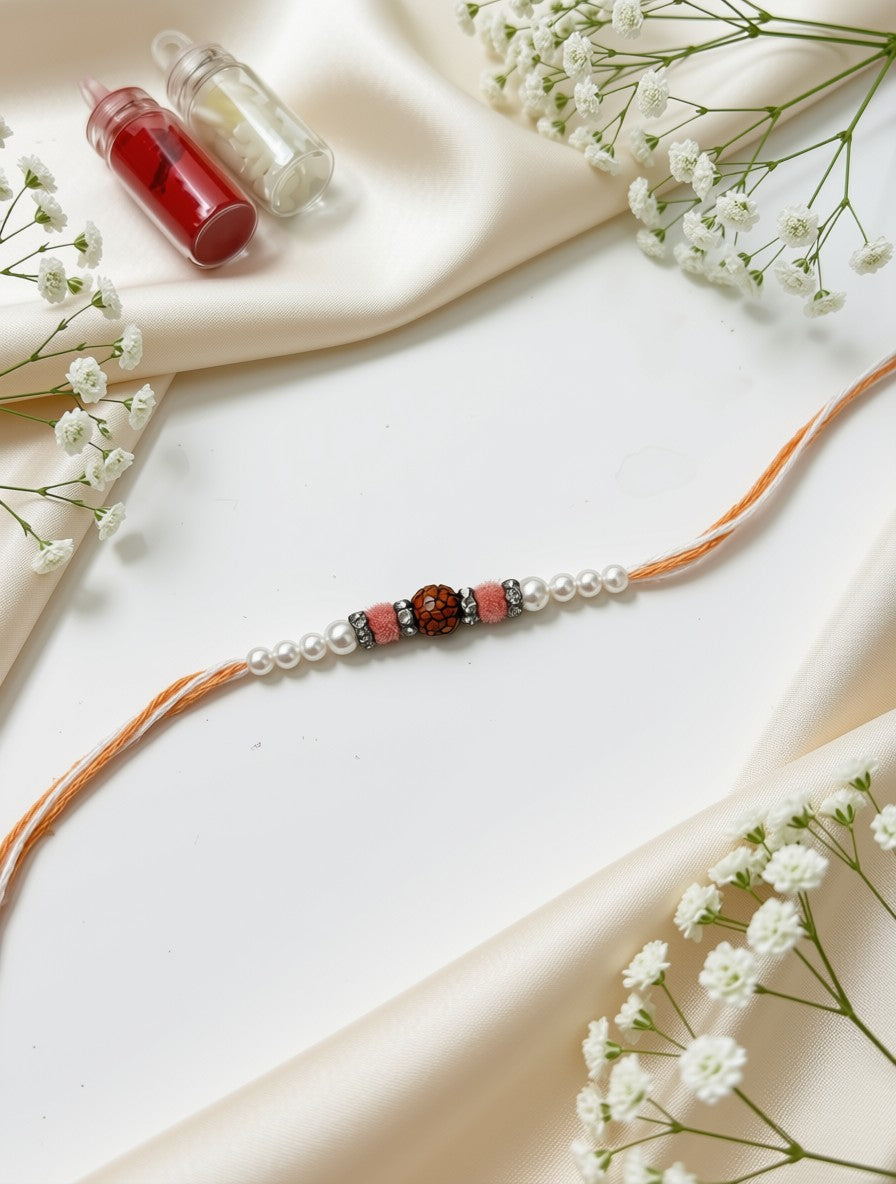 An orange cord bracelet with white faux pearls, pink spacer beads, and a central brown bead, styled on a white surface with cream fabric and baby's breath flowers.
