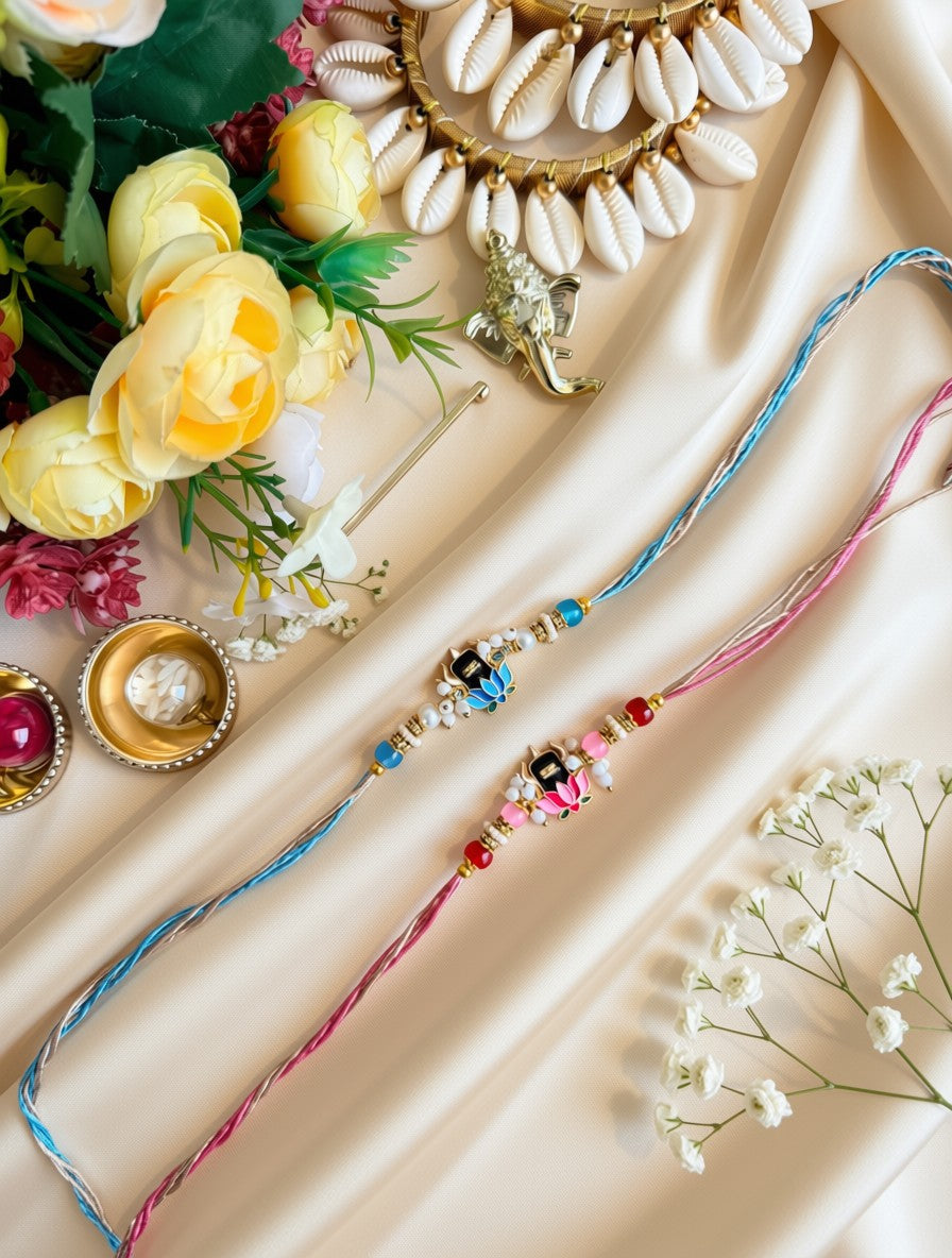 Shivling Blue and Pink Rakhi Two traditional Indian Rakhi beaded wristlets with colorful threads and decorative charms, styled on a beige satin fabric with yellow roses, a gold shell necklace, and small gold bowls.