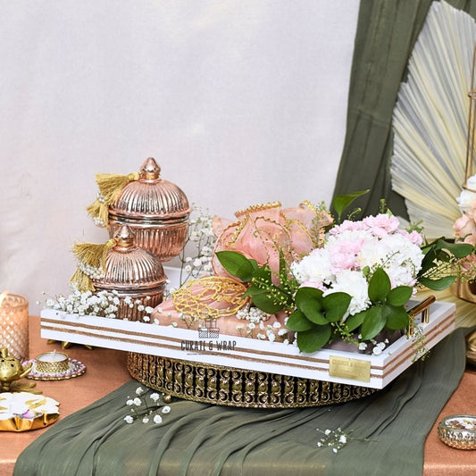 Decorative table setting with gold and pink containers, white flowers, and green leaves on a white tray.
