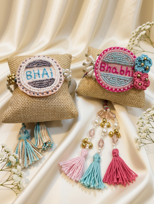 Two handmade burlap wristlets displayed on beige satin fabric with decorative flowers. One wristlet has a circular patch with 'BHAI' in blue, and the other has a patch with 'Bhabhi' in pink, both with colorful tassels and beaded details.