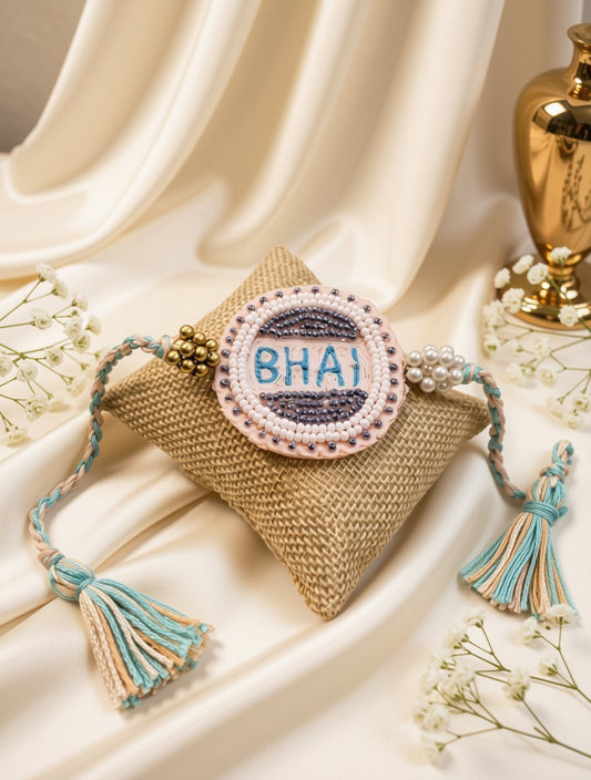 A handmade braided Rakhi with a circular embroidered patch reading 'BHAI' in blue on a navy and lavender striped background, surrounded by a white and pink beaded border. The Rakhi is displayed on a small woven jute cushion, set against a draped cream satin backdrop with decorative flowers and a gold vase.