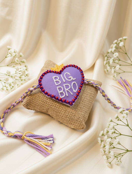 A handmade heart-shaped purple gift tag with 'BIG BRO' embroidered in white, resting on a small burlap pouch. The tag is attached to a purple, lavender, and yellow braided cord with tassels, set against a draped cream satin background with baby's breath flowers.