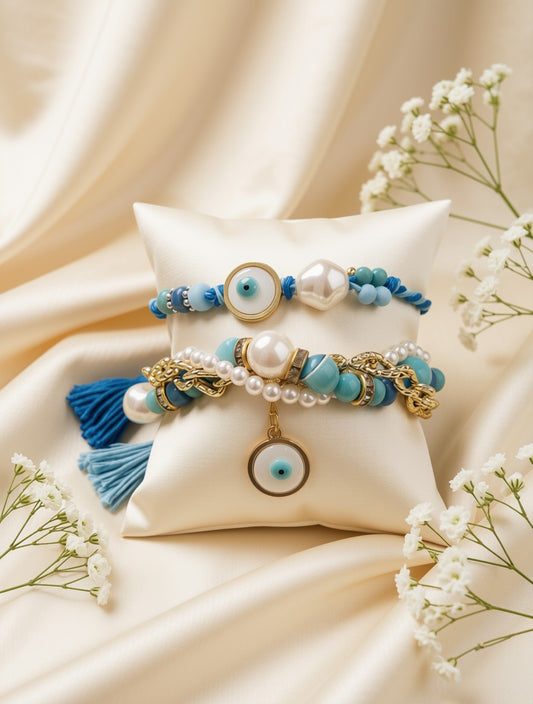 Two bohemian-style beaded bracelets displayed on a cream satin pillow. The bracelets feature light blue and white beads, white faux pearls, gold-toned metal rings, evil eye charms, and one bracelet has a blue tassel.