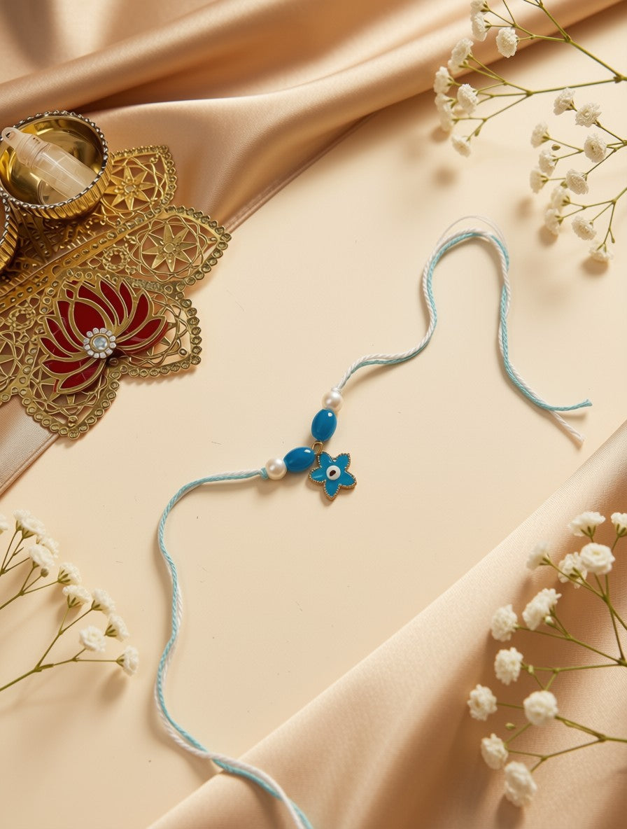A light blue string rakhi with a blue and white evil eye charm, styled on a beige satin background with decorative gold and red jewelry and white baby's breath flowers.