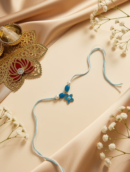 A light blue string rakhi with a blue and white evil eye charm, styled on a beige satin background with decorative gold and red jewelry and white baby's breath flowers.