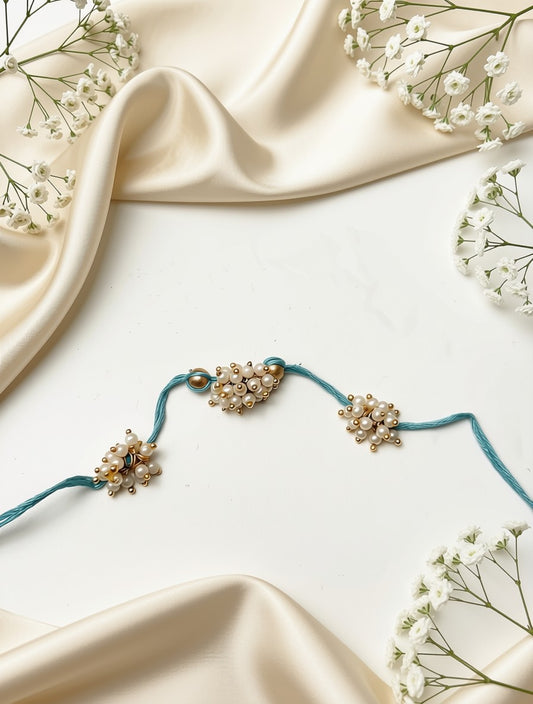 A delicate teal cord jewelry piece with three clusters of small white pearls and gold-toned beads, arranged on a white surface with cream satin fabric and white baby's breath flowers.