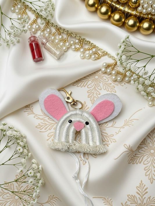 A handmade felt bunny keychain or bag charm with white faux fur trim, pink inner ears, and a pink nose, displayed on draped white satin fabric with pearl strands, gold beads, and baby's breath flowers.