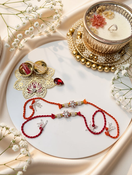 A styled flat-lay of red and gold thread Rakhi bracelets on a white round plate, surrounded by white baby's breath flowers and a gold-toned decorative candle holder on a beige satin background.