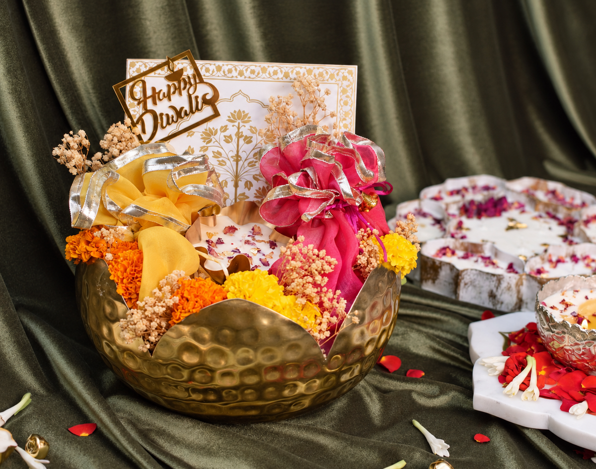 A festive Diwali gift arrangement in a hammered gold bowl, featuring a 'Happy Diwali' card, pink and yellow marigold flowers, dried floral elements, and a silver ribbon. In the background, traditional clay lamps and red rose petals are arranged on a dark green satin drape.