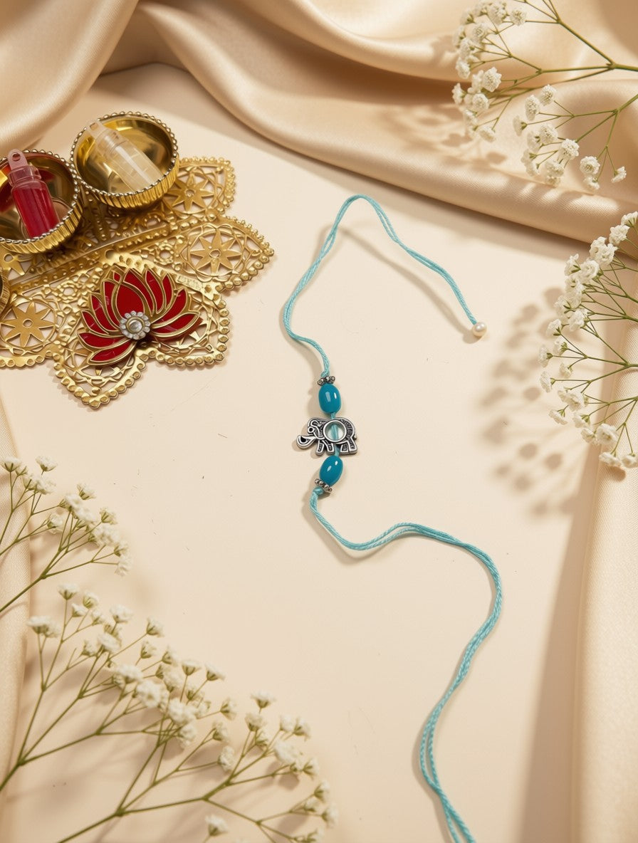 A light blue braided string necklace with a silver-toned elephant charm and turquoise beads, styled on a beige satin fabric with decorative gold and white flowers.
