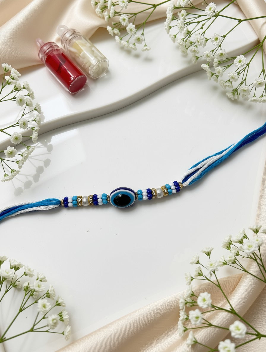 A blue cord bracelet with a central evil eye bead and small blue, white, and gold beads, arranged on a white surface with cream satin fabric and white baby's breath flowers.