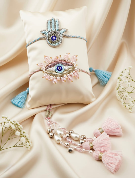 Two beaded Hamsa and eye-themed Hara bracelets displayed on a cream satin pillow, surrounded by draped cream satin fabric, decorative dried flowers, and pink and blue tassels.