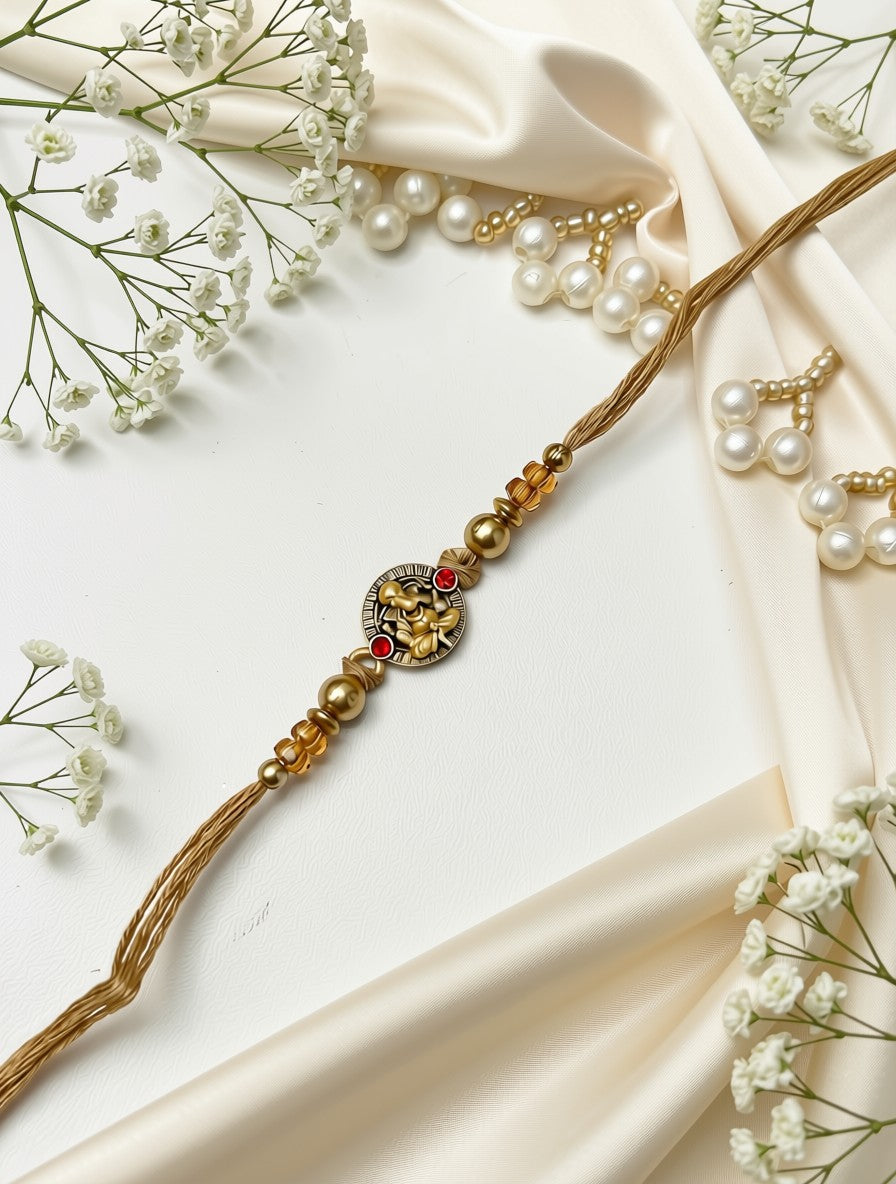 A gold-plated Rakhi with a central elephant charm, red and green stone accents, and gold beads, styled on a cream satin fabric with white pearl earrings and baby's breath flowers.