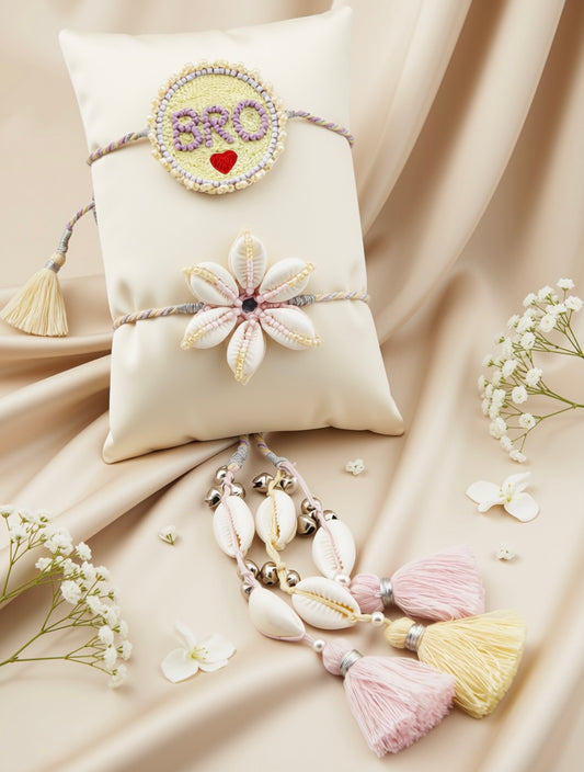 A decorative Rakhi-style wristlet displayed on a cream satin pillow with matching tassel and shell charms, surrounded by small white flowers on draped beige fabric.
