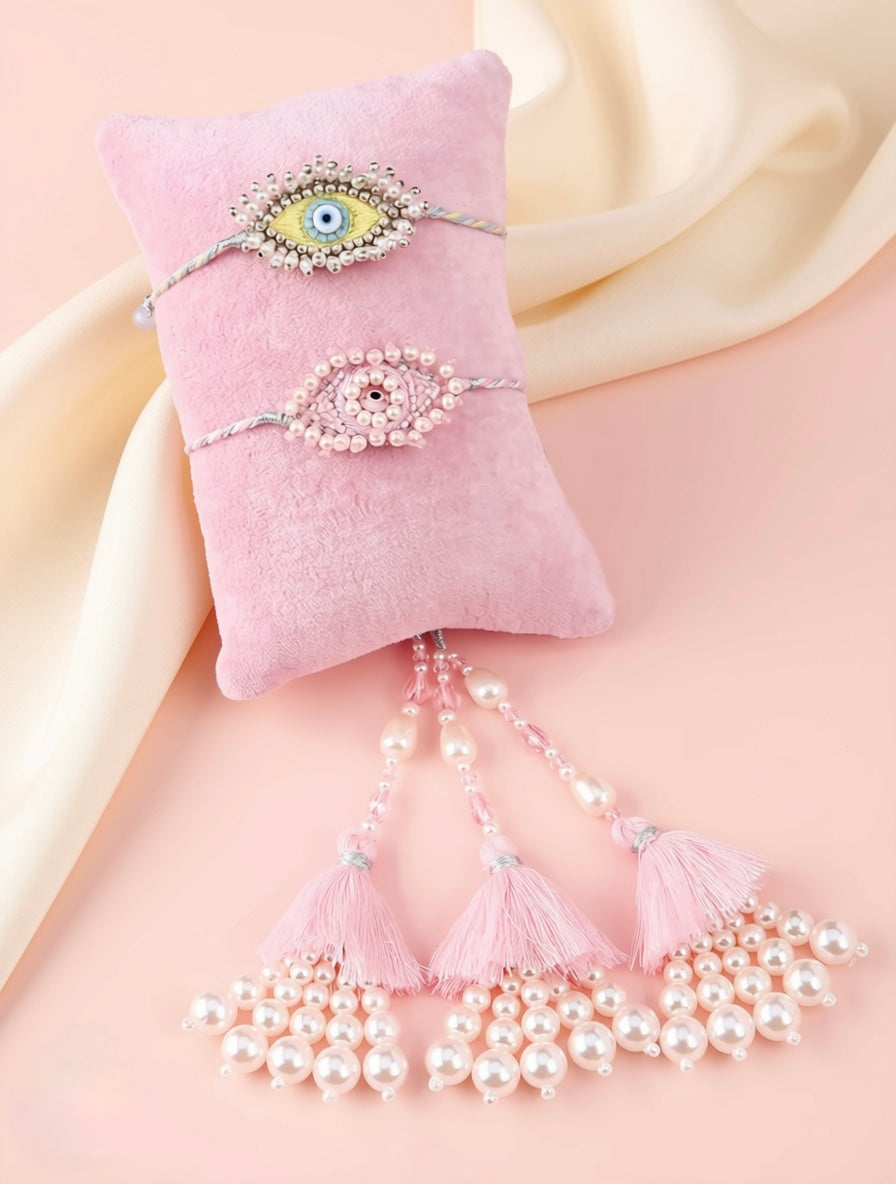 Two pink beaded and pearl-embellished evil eye wristbands with dangling tassels, displayed on a pink velvet pillow against a soft cream satin backdrop.