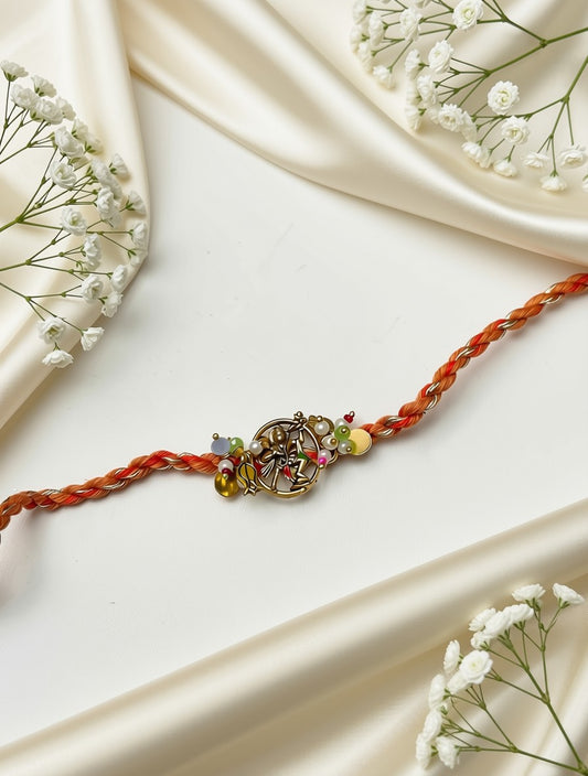 A braided orange-red string bracelet with a central gold-toned decorative charm and small multicolored beads, styled on draped cream satin fabric with white baby's breath flowers.