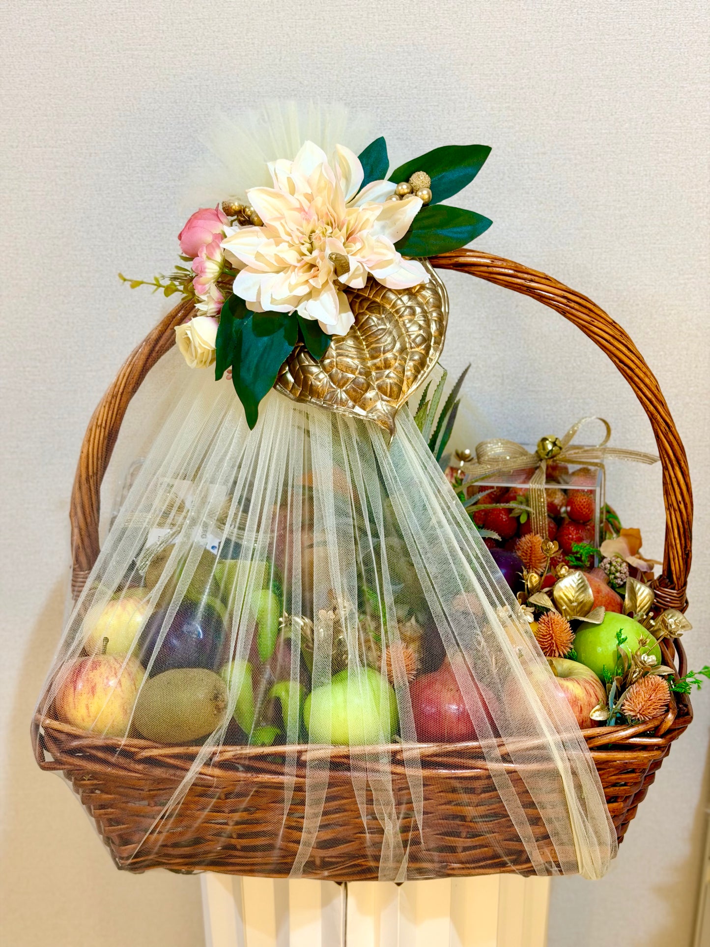 A wicker gift basket filled with assorted fruits, wrapped in sheer white tulle and decorated with artificial flowers, green leaves, and a gold ornament, set against a plain light beige wall.
