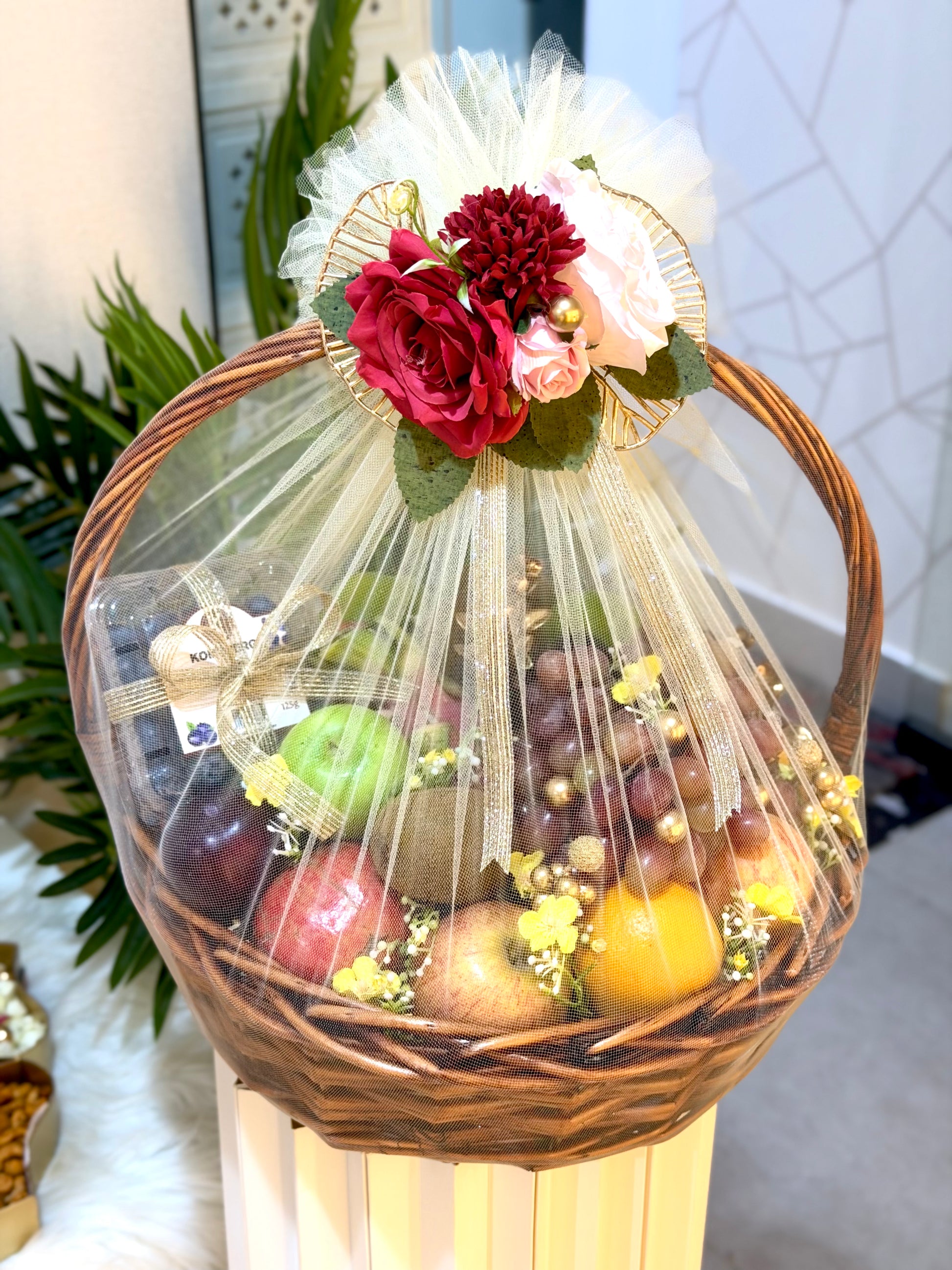 A wicker gift basket filled with assorted fruits, wrapped in clear cellophane and decorated with a white tulle bow, red and pink flowers, and green leaves, displayed on a pedestal in an indoor setting.