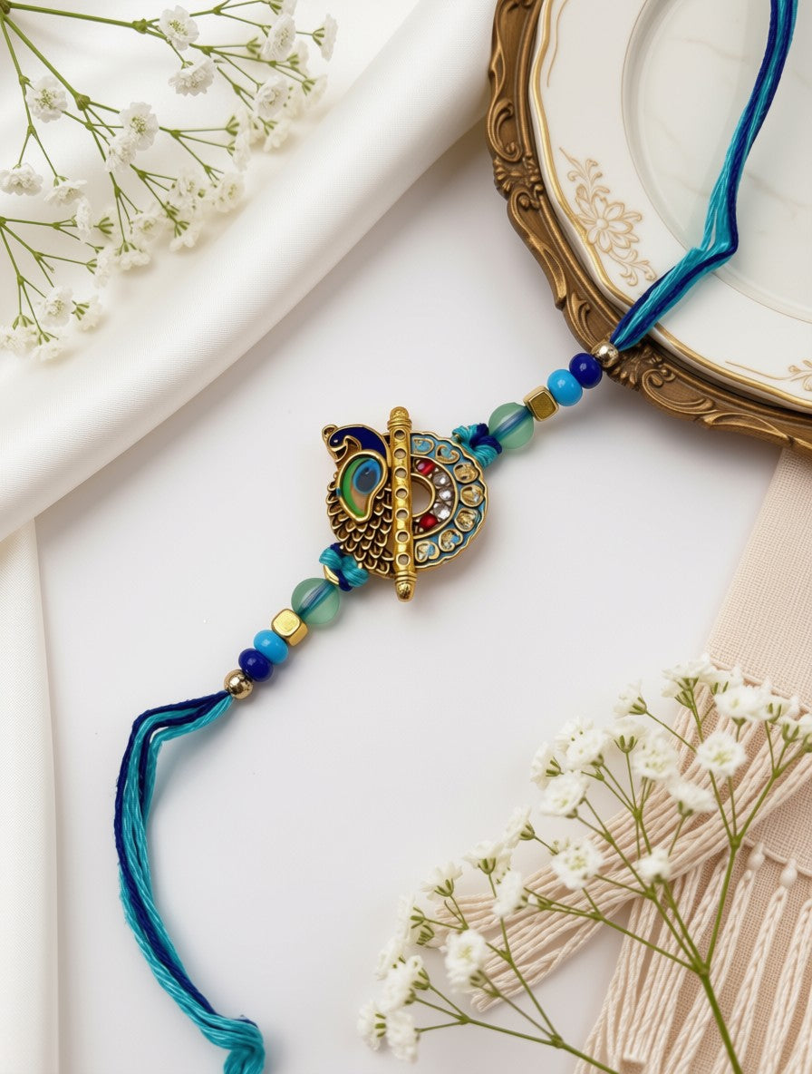 A blue and gold beaded Rakhi with a central peacock ornament, styled on a white fabric background with dried baby's breath flowers and a gold-rimmed plate.