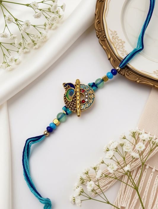 A blue and gold beaded Rakhi with a central peacock ornament, styled on a white fabric background with dried baby's breath flowers and a gold-rimmed plate.