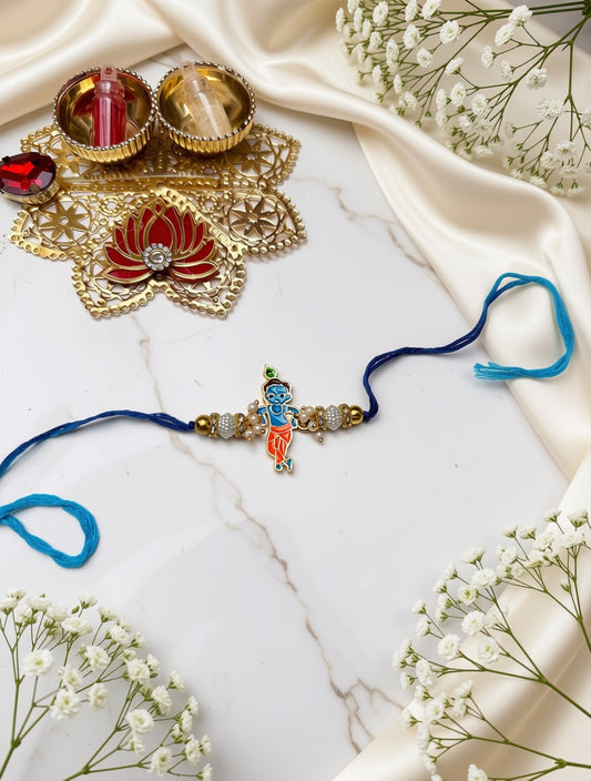 A traditional Indian Rakhi with a colorful Krishna pendant, featuring a navy blue cord with a blue loop and a gold-toned beaded bracelet, styled on a white marble surface with satin fabric, diyas, and baby's breath flowers.