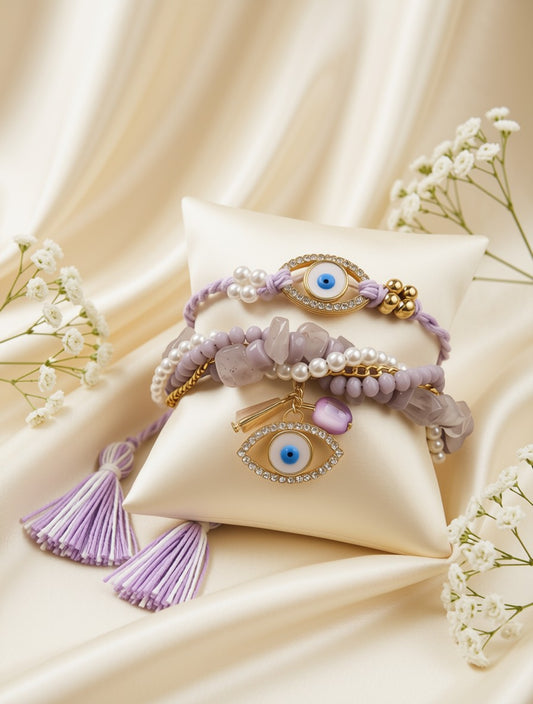 A stack of three purple and lavender bracelets displayed on a cream satin pillow. The bracelets feature evil eye charms with blue centers, white faux pearls, lavender beads, purple crystal chips, and gold-toned metal accents.