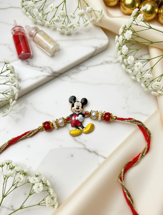 A Mickey Mouse-themed rakhi bracelet with red and gold thread, red and white beads, and a central Mickey charm, styled on a white marble surface with baby's breath flowers and gold ornaments.