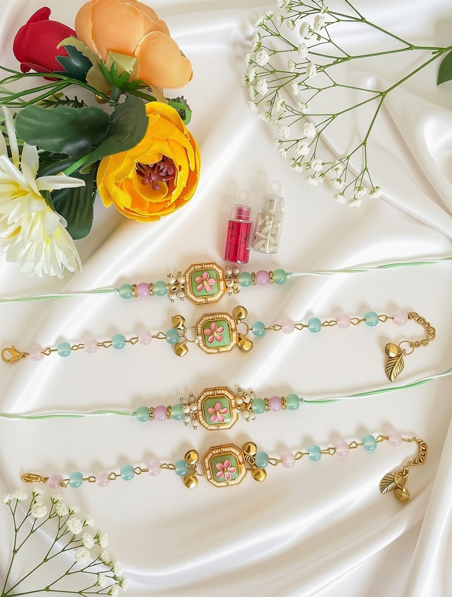 Three traditional Indian Rakhi bracelets with gold-plated borders, pastel green and pink beads, and central floral motifs, arranged on a white satin fabric with decorative flowers and small bottles.