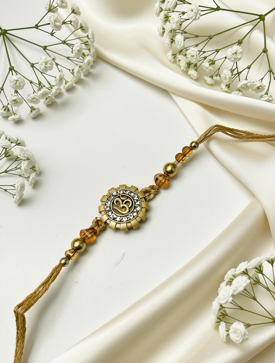 A gold-toned beaded Rakhi with a central Om symbol charm, laid diagonally on draped cream satin fabric with small white baby's breath flowers around it.