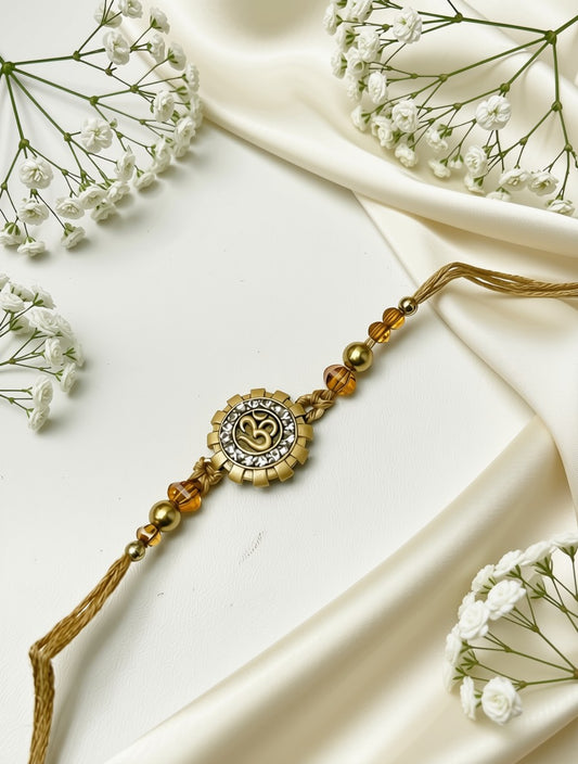 A gold-toned beaded Rakhi with a central Om symbol charm, laid diagonally on draped cream satin fabric with small white baby's breath flowers around it.