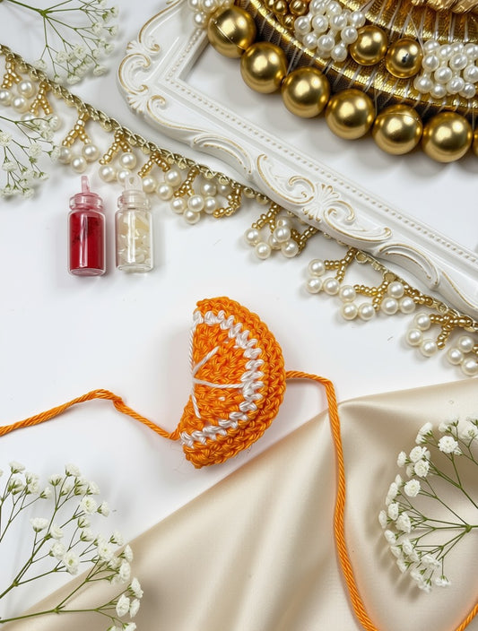 A crocheted orange slice ornament or decorative piece in orange and white yarn, styled on a white surface with pearls, gold beads, baby's breath flowers, and small bottles of red and clear liquid.