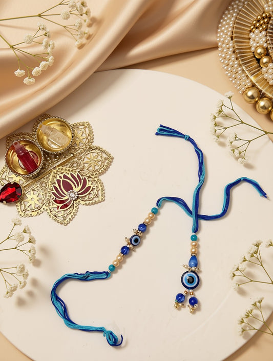 A blue beaded string accessory with evil eye motifs and tassels, arranged on a white oval plate with decorative gold jewelry, pearl beads, and dried flowers around it.