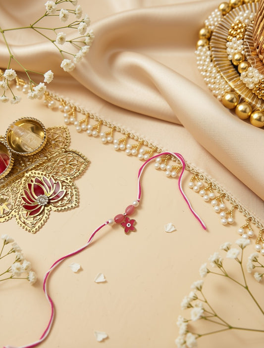 A pink Rakhi with a small red flower charm and white beads, laid out on a beige satin fabric alongside gold foil borders, pearl trim, and baby's breath flowers.