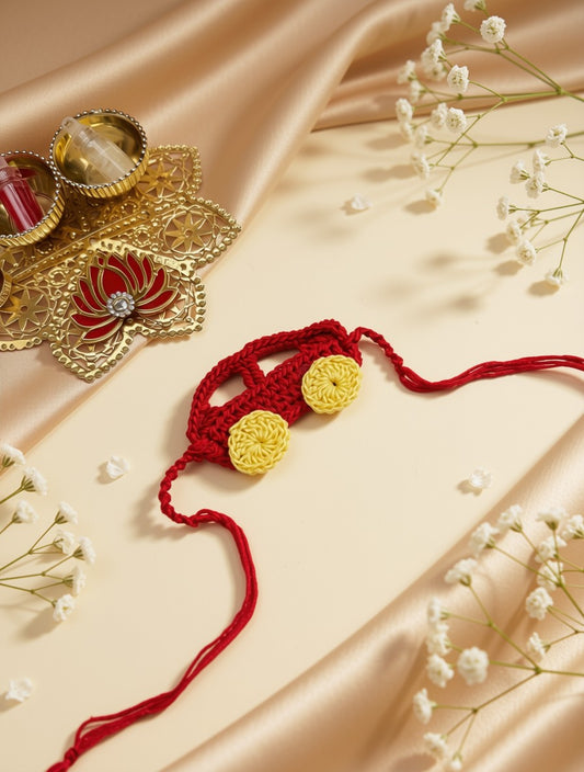 A red crocheted Rakhi with yellow circular accents, laid on a beige satin fabric with white baby's breath flowers and a decorative gold diya in the background.