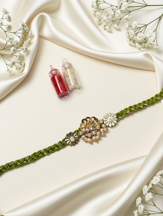 A green braided bracelet with a central floral charm featuring gold, silver, and white elements, styled on a cream satin background with small decorative bottles and baby's breath flowers.