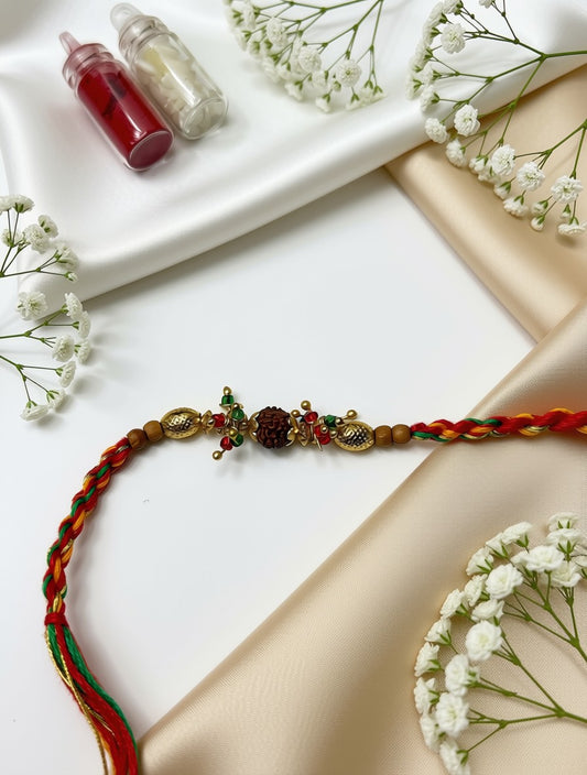 A traditional Indian Rakhi with braided red, orange, and green threads, gold-toned beads, and a central dark red and green ornament, displayed on a white and beige satin background with baby's breath flowers and small glass bottles.