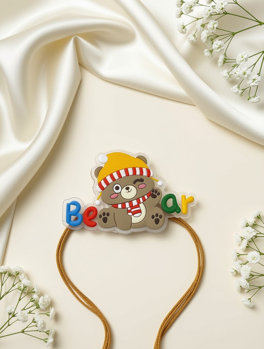 A headband with a cartoon bear wearing a yellow hat and red-and-white striped scarf, alongside colorful letters spelling 'Bear'. The headband has tan elastic cords and is styled on a cream satin fabric background with small white flowers.