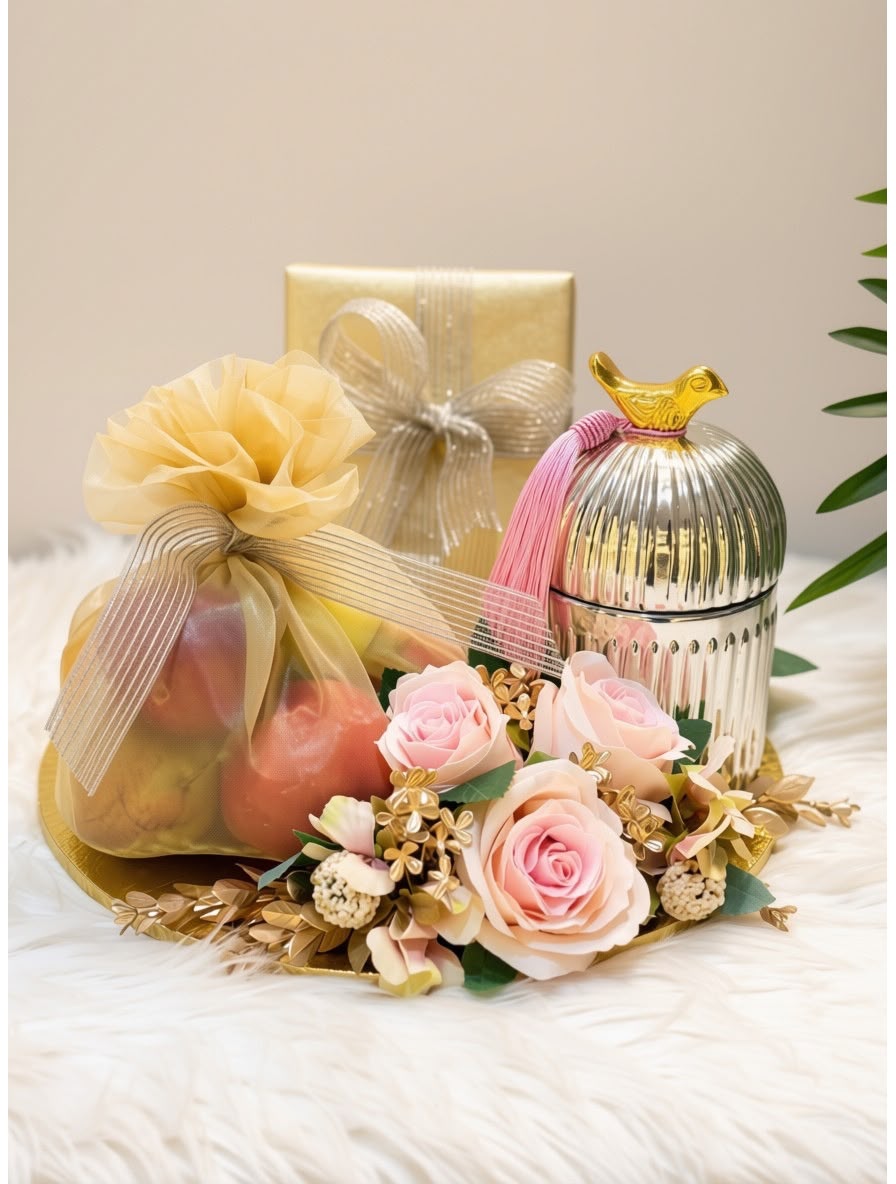 A styled gift arrangement featuring a sheer mesh bag filled with red apples tied with a cream ribbon, a metallic silver ribbed lidded container with a pink ribbon and gold bird topper, a gold gift box in the back, pink roses, and dried floral accents on a white faux-fur surface.